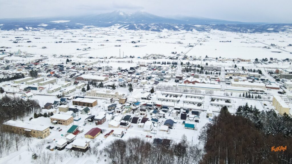 中富良野町丘町の空撮。雪に覆われた街並みと十勝岳連峰のパノラマビュー
